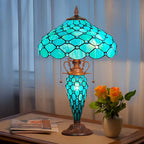 Decorative table lamp with stained glass shade on a table next to a vase of flowers.