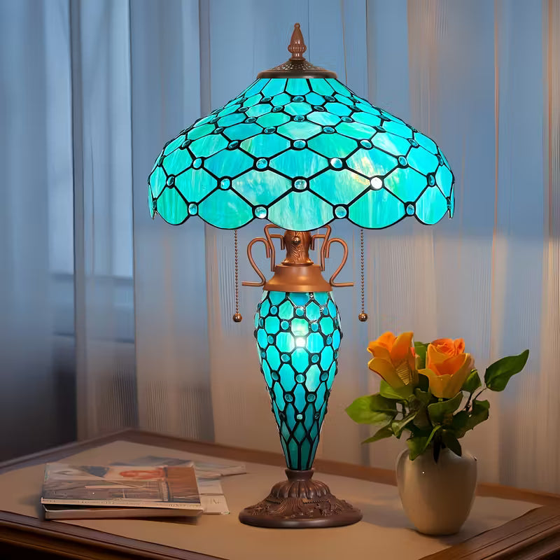 Decorative table lamp with stained glass shade on a table next to a vase of flowers.