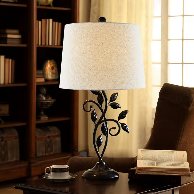 Decorative table lamp with black metal base and white shade on a table next to a book and cup.