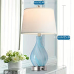 Table lamp with blue glass base and white shade on a surface with a vase of flowers in the background.