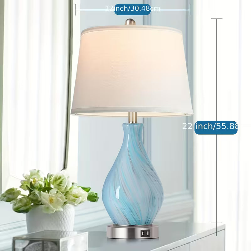 Table lamp with blue glass base and white shade on a surface with a vase of flowers in the background.